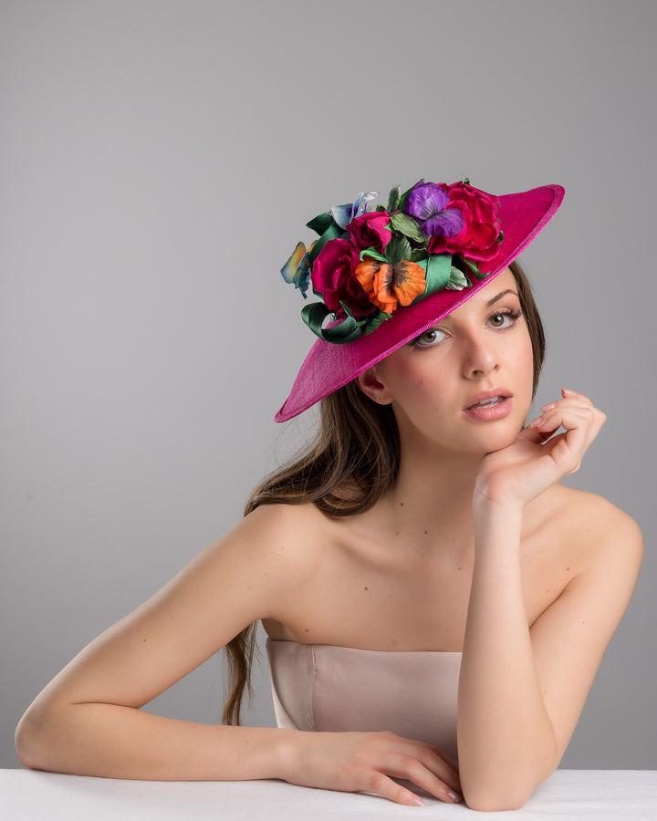 The female model is posing, wearing Jalen the medium sized, hot pink medium teardrop saucer decorated with a bouquet of eye-catching pink roses, multi-coloured violets and emerald leaves. Designed and made by Merve Bayindir Millinery.
