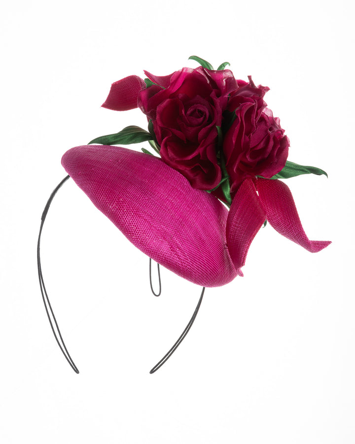 Hot pink sinamay fascinator with a sculpted asymmetrical base, topped with deep red rose blooms and green leaves, mounted on a slim black headband. Designed and made by Merve Bayindir Millinery.
