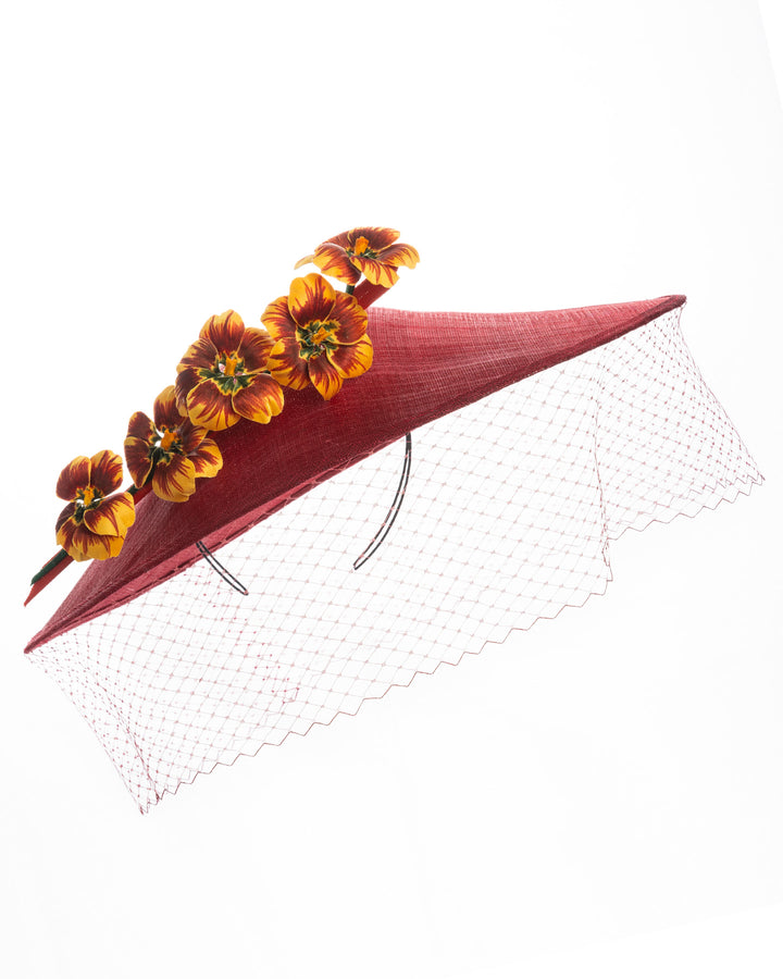 Deep red sinamay wide-brim fascinator with a sculptural pointed shape, decorated with orange and burgundy pansy flowers and a matching red birdcage veil. Designed and made by Merve Bayindir Millinery.

