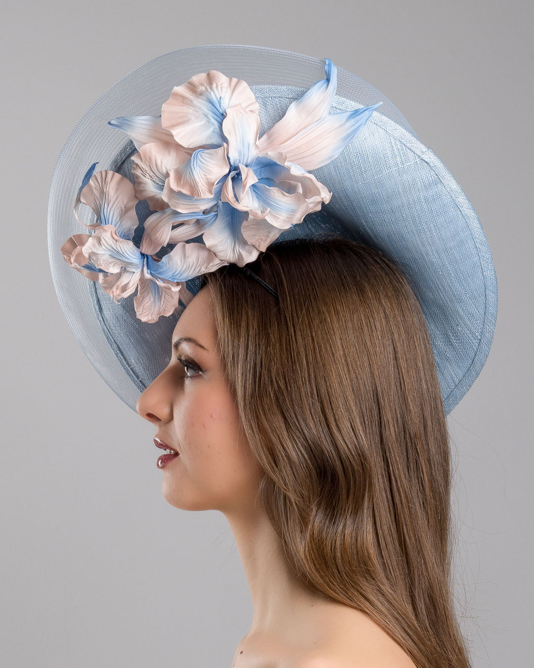 The model is showcasing the left side close up of the medium sized, side saucer shaped light blue headpiece that blends different styles into one. Almost vertical construction of this splendid design softens with a crinoline curve on the left side and features light pink and light blue shaded irises underneath. Designed and made by Merve Bayindir Millinery