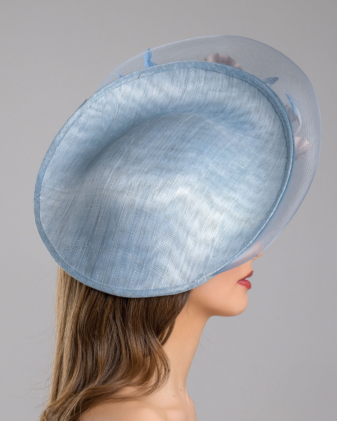 The model is showcasing the right side close up of the medium sized, side saucer shaped light blue headpiece that blends different styles into one. Almost vertical construction of this splendid design softens with a crinoline curve on the left side and features light pink and light blue shaded irises underneath. Designed and made by Merve Bayindir Millinery