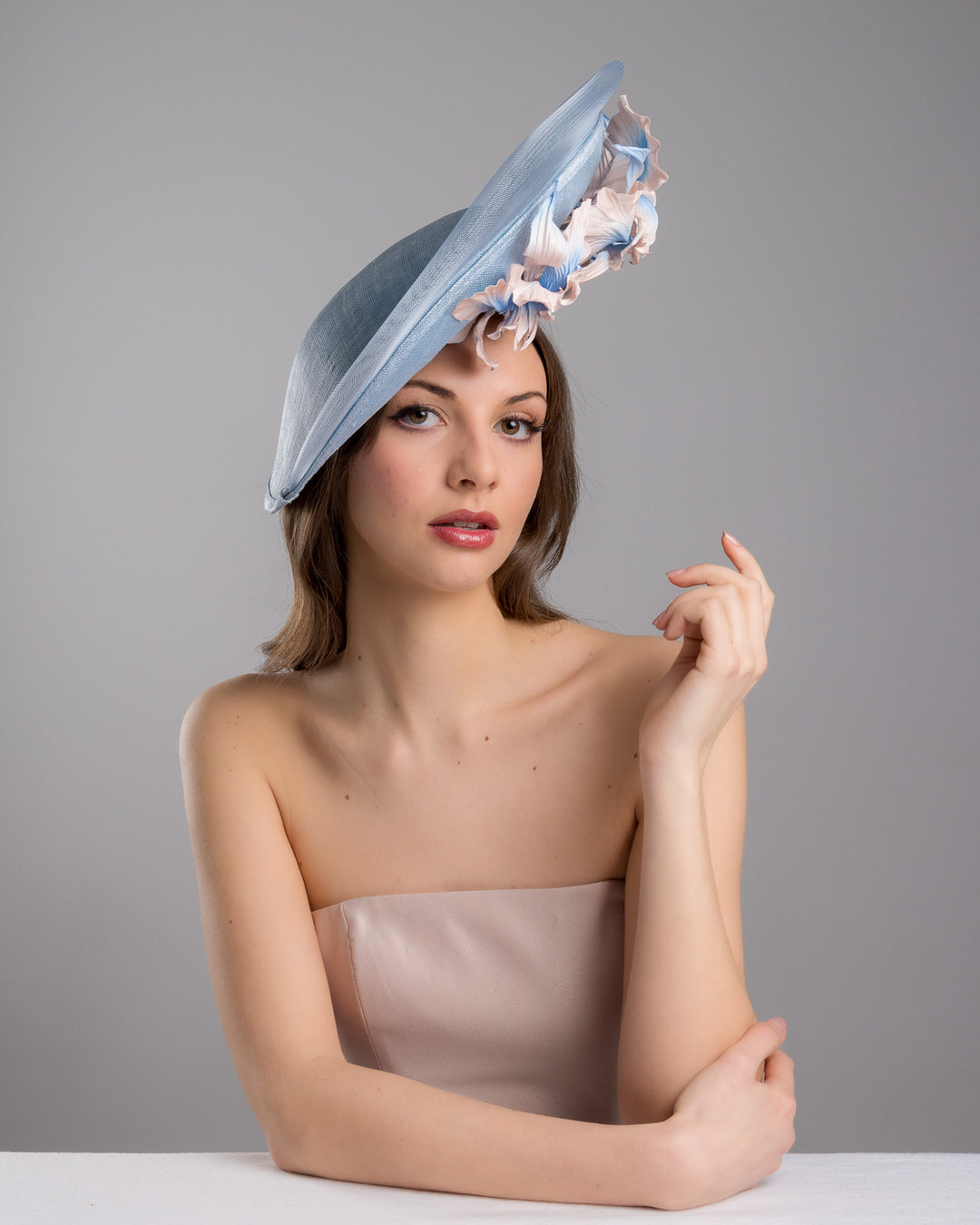 The female model is posing, wearing Jessie,the medium sized, side saucer shaped light blue headpiece that blends different styles into one. Almost vertical construction of this splendid design softens with a crinoline curve on the left side and features light pink and light blue shaded irises underneath. Designed and made by Merve Bayindir Millinery