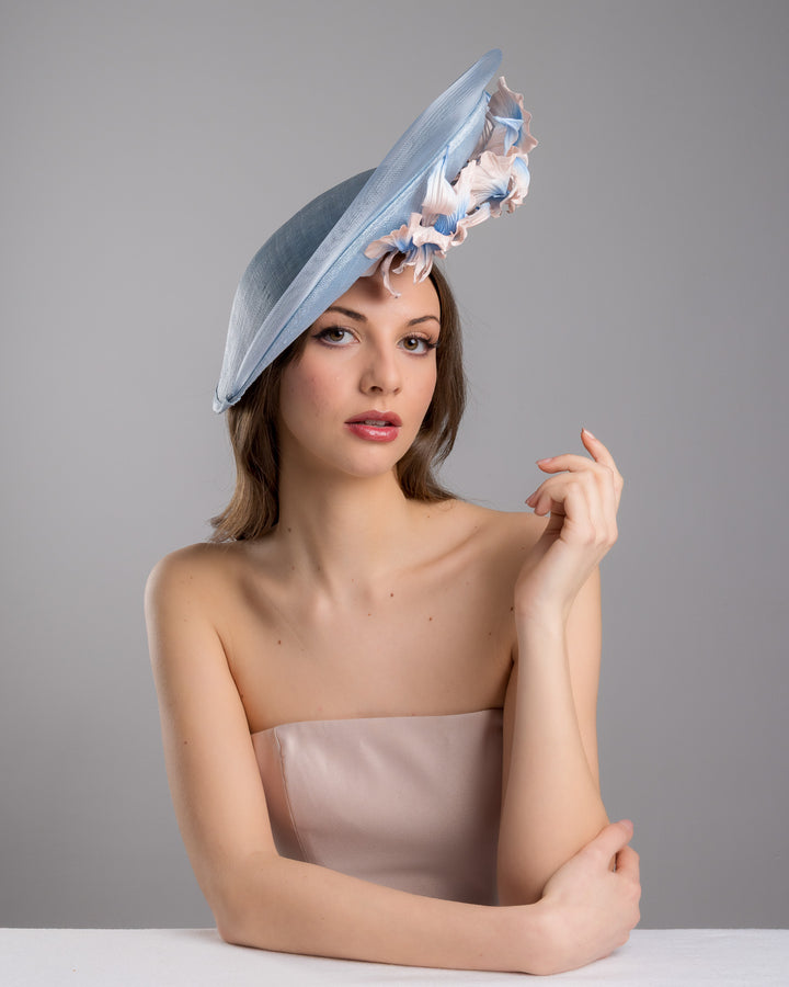 The female model is posing, wearing Jessie,the medium sized, side saucer shaped light blue headpiece that blends different styles into one. Almost vertical construction of this splendid design softens with a crinoline curve on the left side and features light pink and light blue shaded irises underneath. Designed and made by Merve Bayindir Millinery