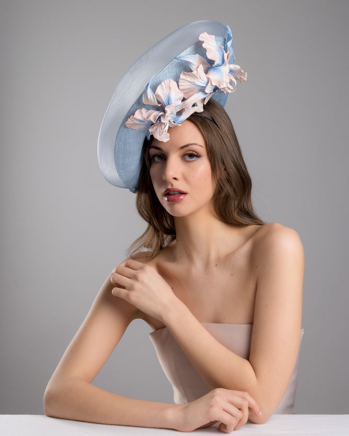The female model is posing, wearing Jessie,the medium sized, side saucer shaped light blue headpiece that blends different styles into one. Almost vertical construction of this splendid design softens with a crinoline curve on the left side and features light pink and light blue shaded irises underneath. Designed and made by Merve Bayindir Millinery