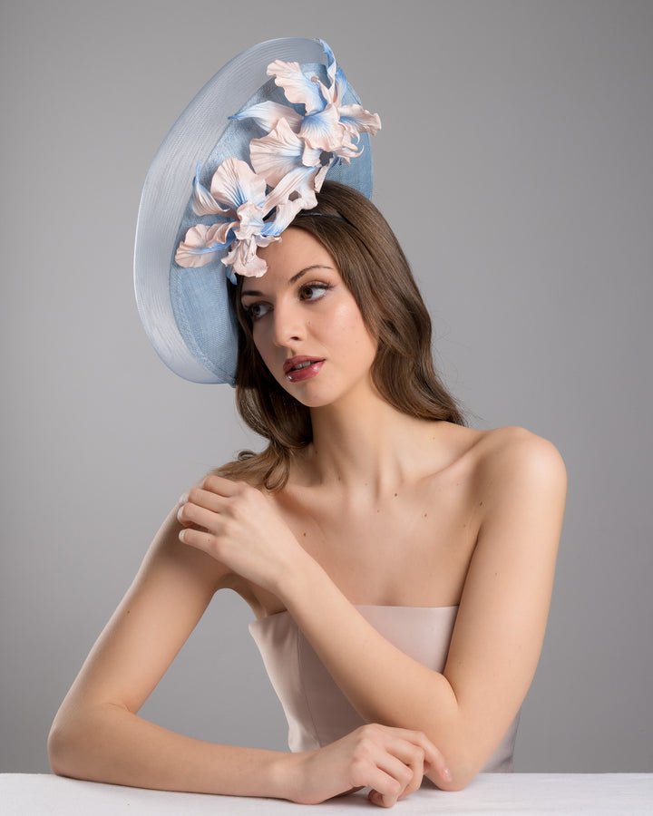 The female model is posing, wearing Jessie,the medium sized, side saucer shaped light blue headpiece that blends different styles into one. Almost vertical construction of this splendid design softens with a crinoline curve on the left side and features light pink and light blue shaded irises underneath. Designed and made by Merve Bayindir Millinery