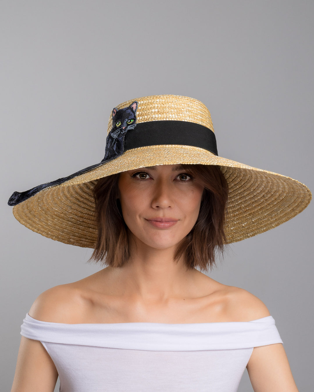 The model is showcasing the front side close up of the natural woven wide brim straw boater hat with a wide brim and black grosgrain ribbon band, featuring a large black cat appliqué with green eyes draped over the crown and brim. Design and made by Merve Bayindir Millinery.
