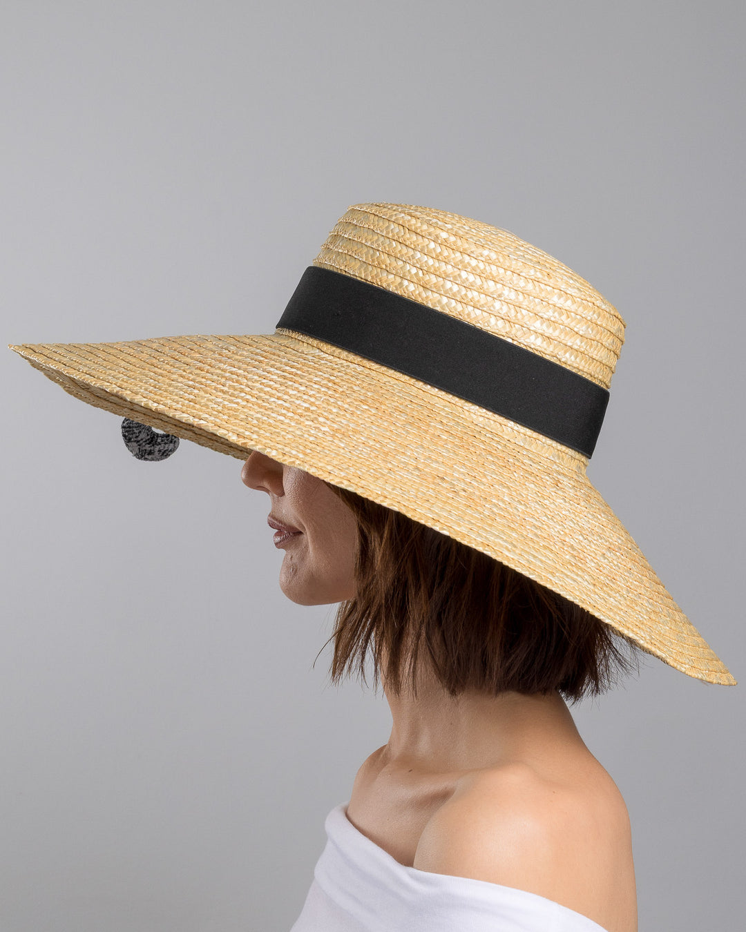 The model is showcasing the left side close up of the natural woven wide brim straw boater hat with a wide brim and black grosgrain ribbon band, featuring a large black cat appliqué with green eyes draped over the crown and brim. Design and made by Merve Bayindir Millinery.