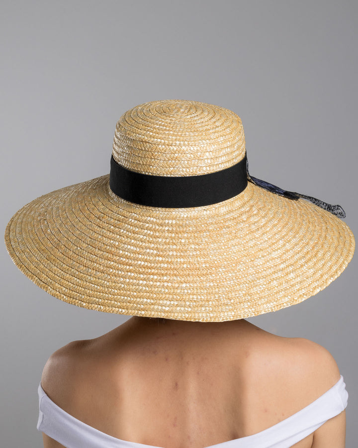 The model is showcasing the back side close up of the natural woven wide brim straw boater hat with a wide brim and black grosgrain ribbon band, featuring a large black cat appliqué with green eyes draped over the crown and brim. Design and made by Merve Bayindir Millinery.