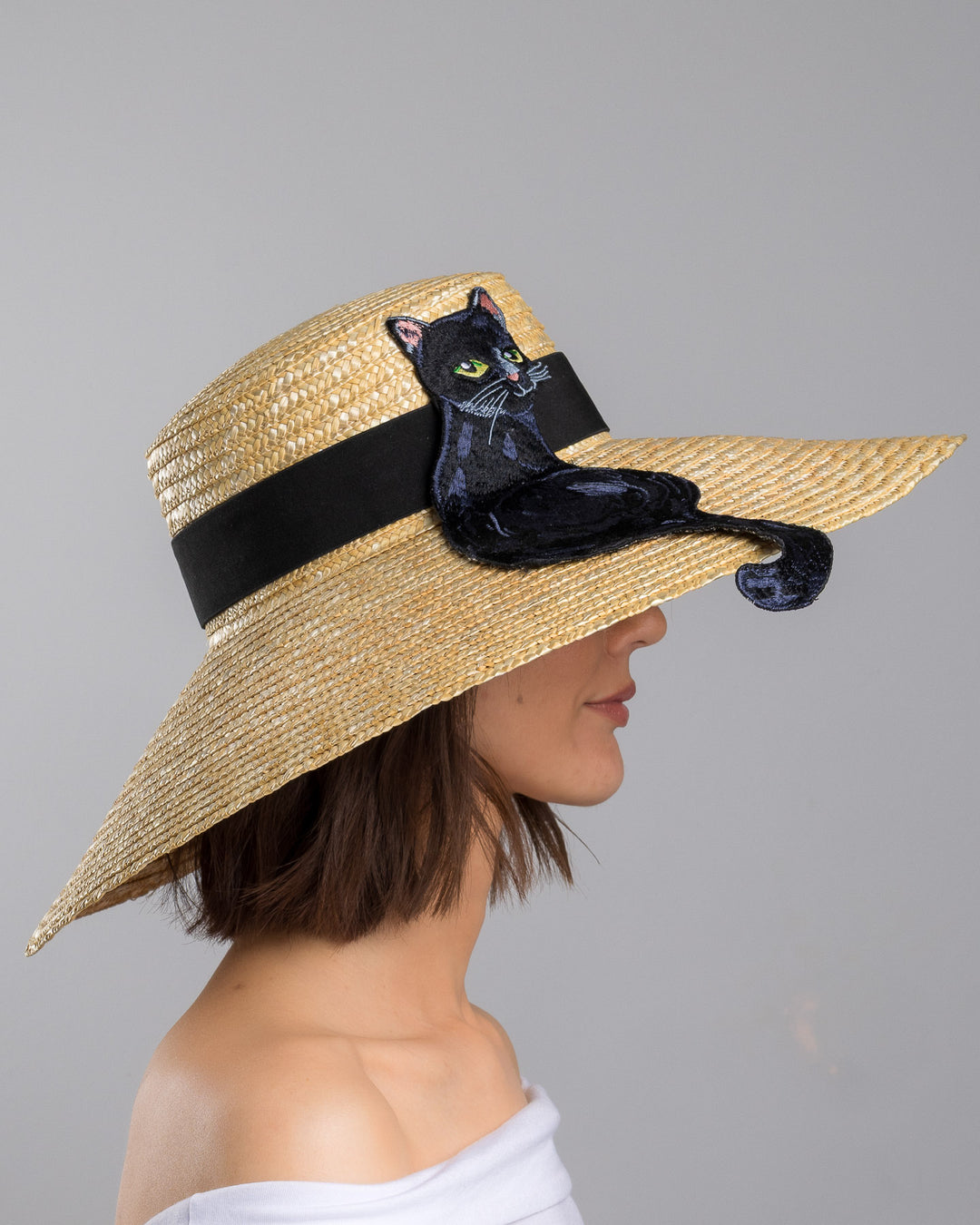 The model is showcasing the right side close up of the natural woven wide brim straw boater hat with a wide brim and black grosgrain ribbon band, featuring a large black cat appliqué with green eyes draped over the crown and brim. Design and made by Merve Bayindir Millinery.