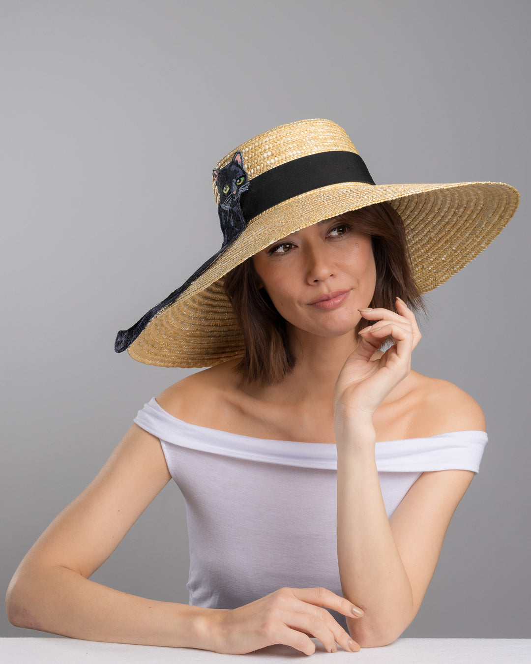 The female model is posing, wearing Kael, the natural woven wide brim straw boater hat with a wide brim and black grosgrain ribbon band, featuring a large black cat appliqué with green eyes draped over the crown and brim. Design and made by Merve Bayindir Millinery.
