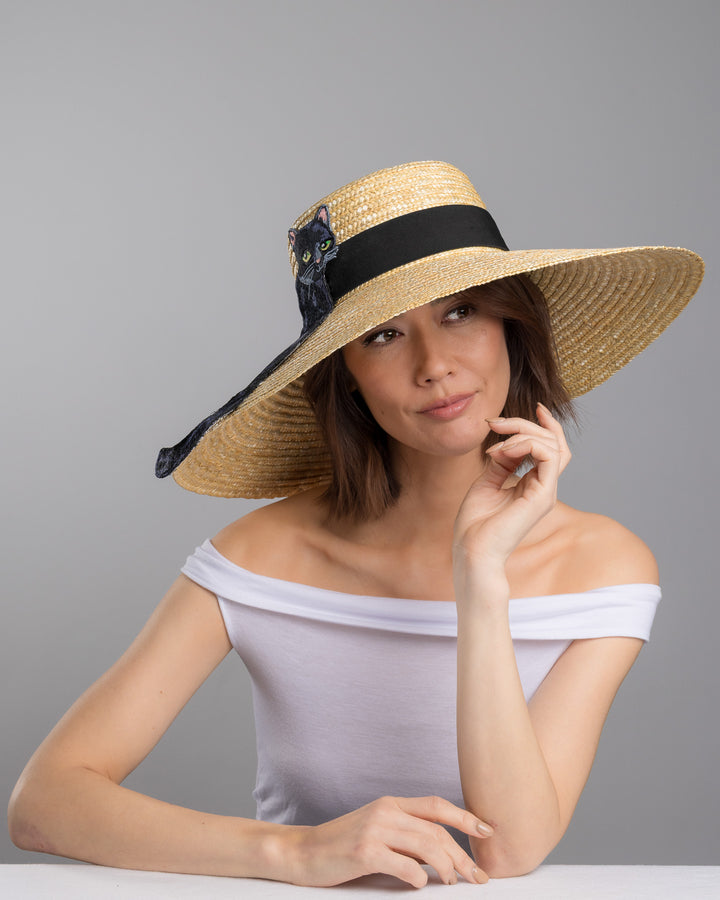 The female model is posing, wearing Kael, the natural woven wide brim straw boater hat with a wide brim and black grosgrain ribbon band, featuring a large black cat appliqué with green eyes draped over the crown and brim. Design and made by Merve Bayindir Millinery.