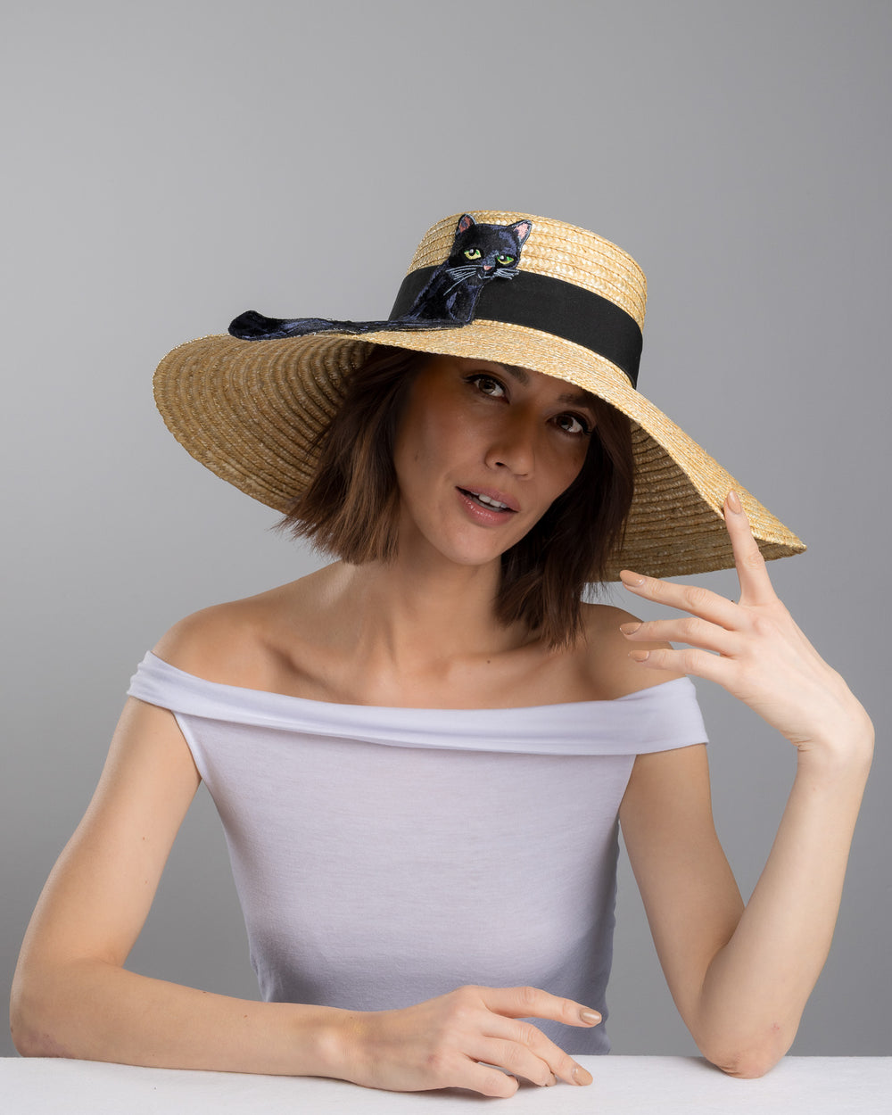 The female model is posing, wearing Kael, the natural woven wide brim straw boater hat with a wide brim and black grosgrain ribbon band, featuring a large black cat appliqué with green eyes draped over the crown and brim. Design and made by Merve Bayindir Millinery.