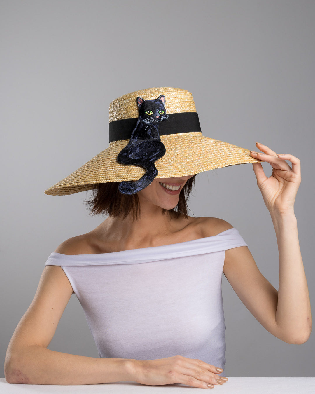 The female model is posing, wearing Kael, the natural woven wide brim straw boater hat with a wide brim and black grosgrain ribbon band, featuring a large black cat appliqué with green eyes draped over the crown and brim. Design and made by Merve Bayindir Millinery.