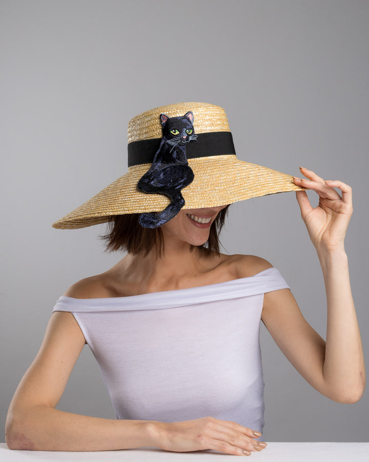 The female model is posing, wearing Kael, the natural woven wide brim straw boater hat with a wide brim and black grosgrain ribbon band, featuring a large black cat appliqué with green eyes draped over the crown and brim. Design and made by Merve Bayindir Millinery.
