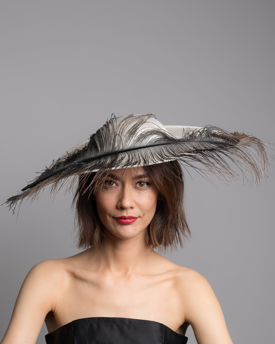 The model is showcasing the front side close up of the single long, curved feather in shades of black, with fine wispy strands and a visible central quill, placed against a plain white background. Designed and made by Merve Bayindir Millinery.