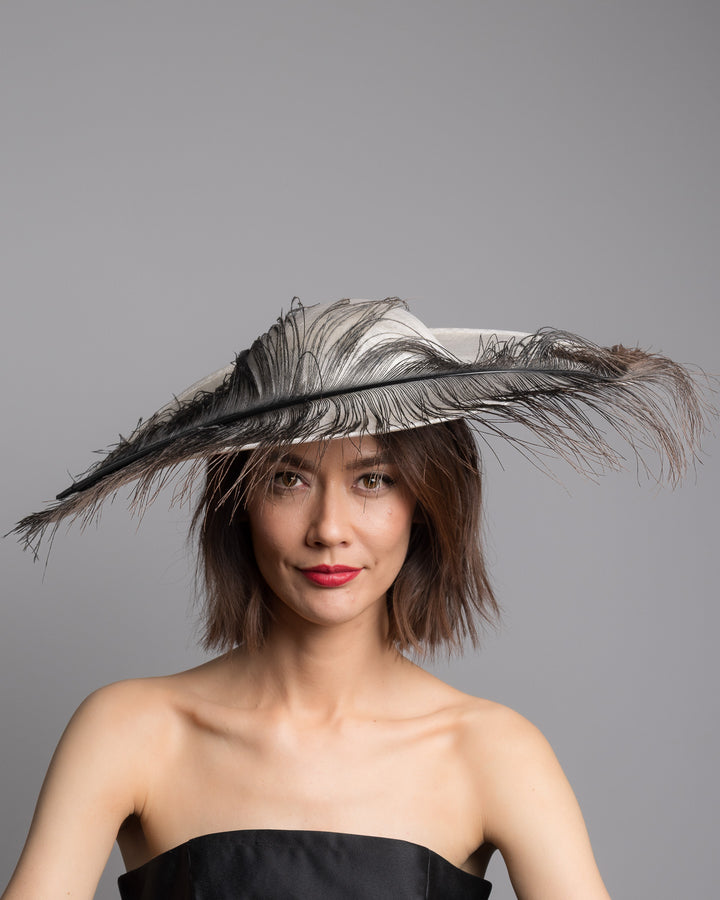 The model is showcasing the front side close up of the single long, curved feather in shades of black, with fine wispy strands and a visible central quill, placed against a plain white background. Designed and made by Merve Bayindir Millinery.