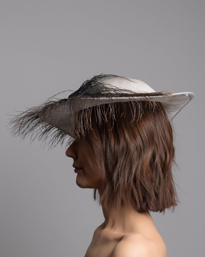 The model is showcasing the left side close up of the single long, curved feather in shades of black, with fine wispy strands and a visible central quill, placed against a plain white background. Designed and made by Merve Bayindir Millinery.