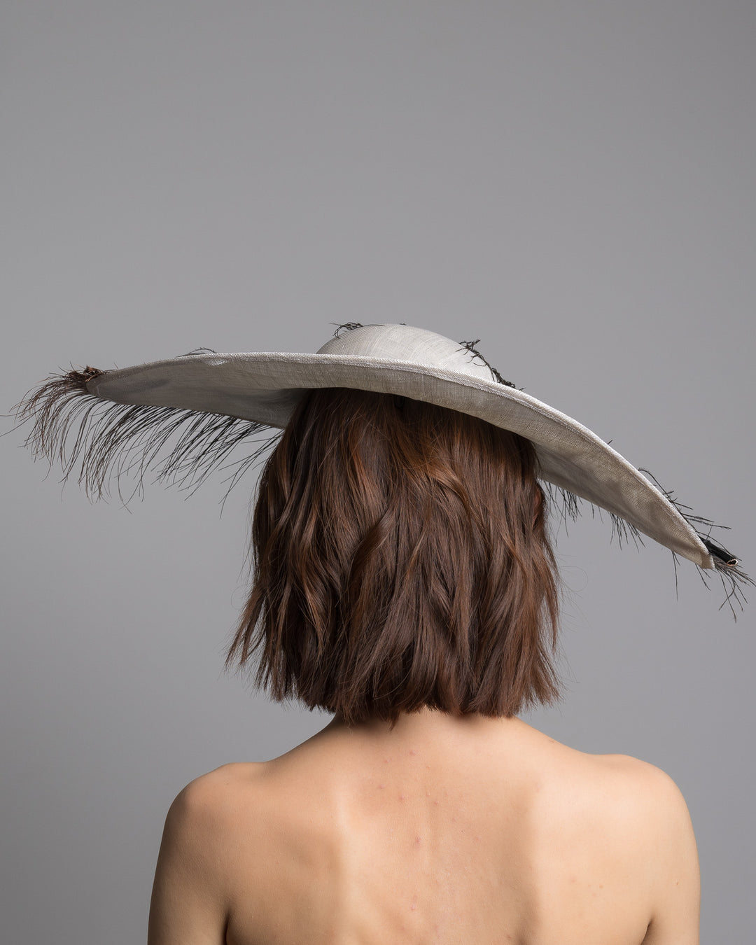 The model is showcasing the back side close up the single long, curved feather in shades of black, with fine wispy strands and a visible central quill, placed against a plain white background. Designed and made by Merve Bayindir Millinery.