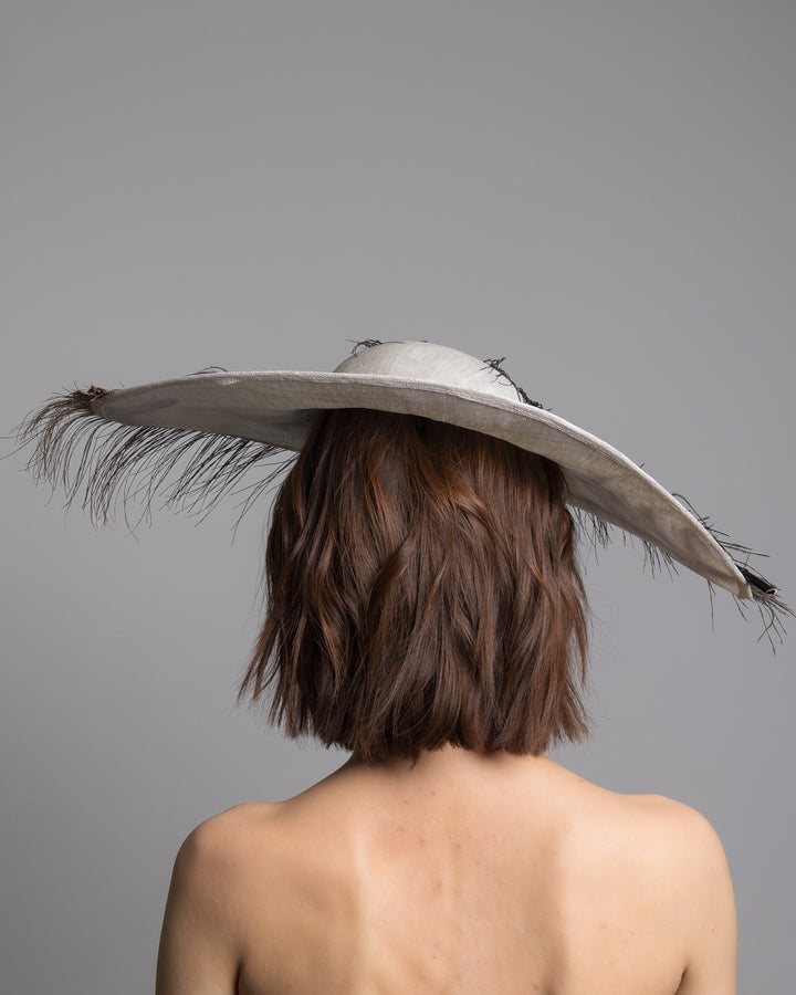 The model is showcasing the back side close up the single long, curved feather in shades of black, with fine wispy strands and a visible central quill, placed against a plain white background. Designed and made by Merve Bayindir Millinery.