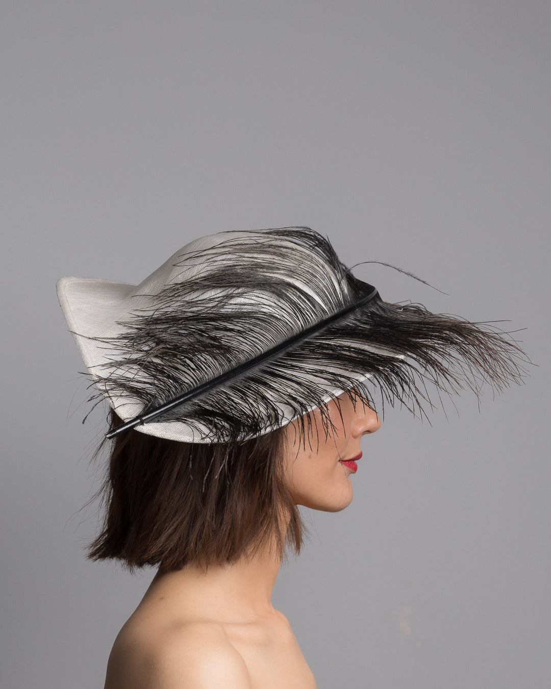 The model is showcasing the right side close up of the single long, curved feather in shades of black, with fine wispy strands and a visible central quill, placed against a plain white background. Designed and made by Merve Bayindir Millinery.
