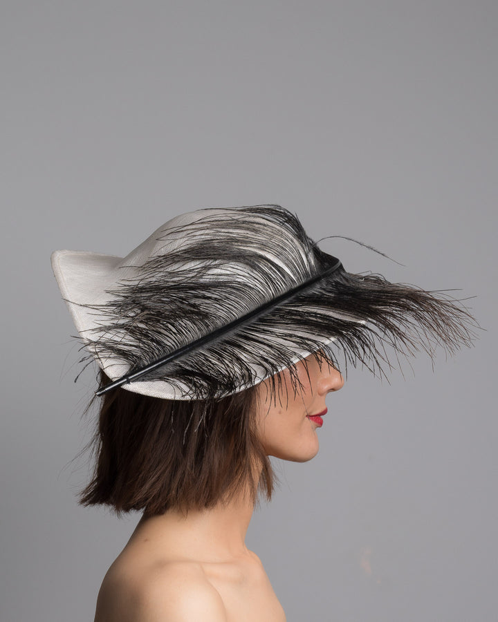 The model is showcasing the right side close up of the single long, curved feather in shades of black, with fine wispy strands and a visible central quill, placed against a plain white background. Designed and made by Merve Bayindir Millinery.