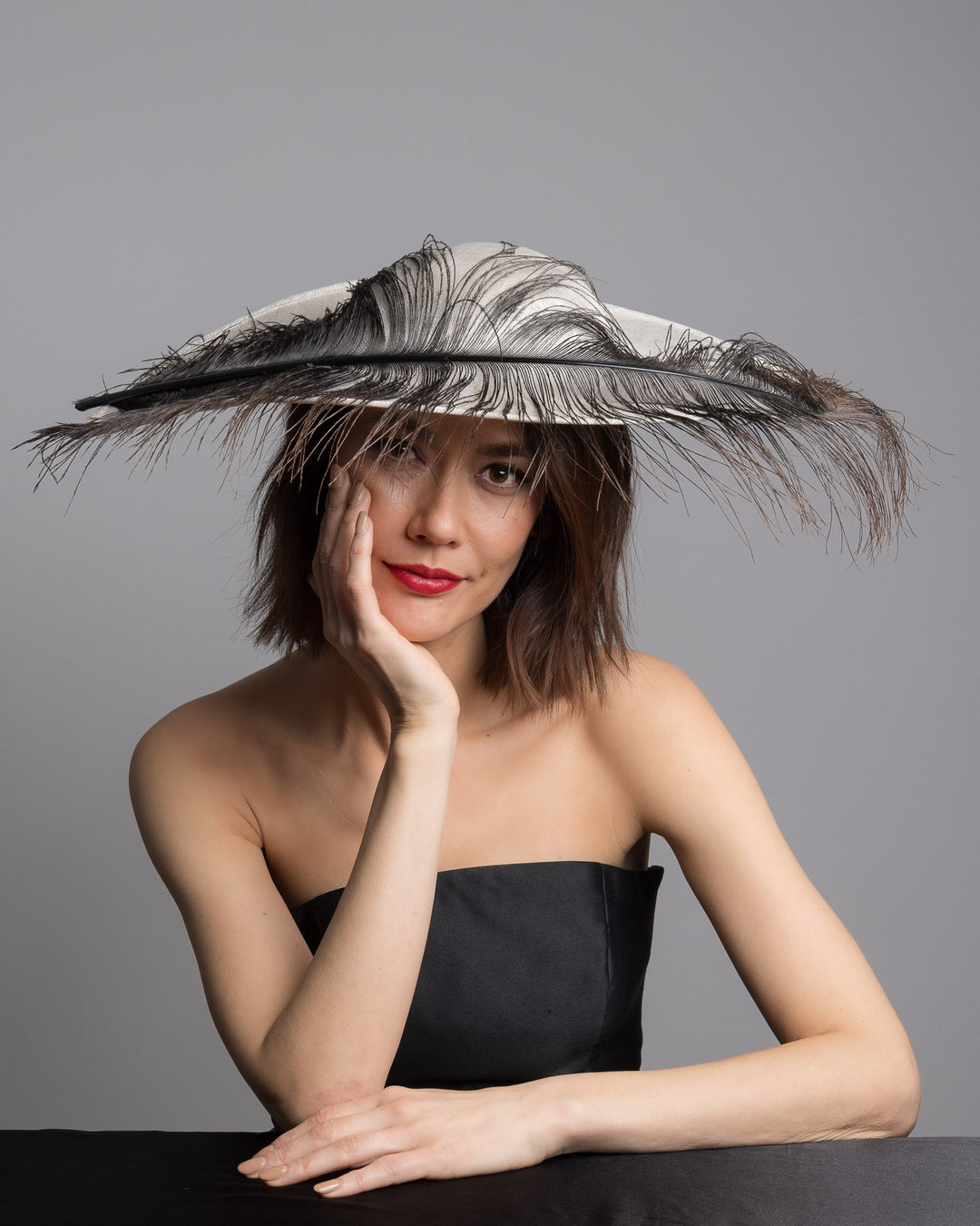 The female model is posing, wearing Kaul, single long, curved feather in shades of black, with fine wispy strands and a visible central quill, placed against a plain white background. Designed and made by Merve Bayindir Millinery