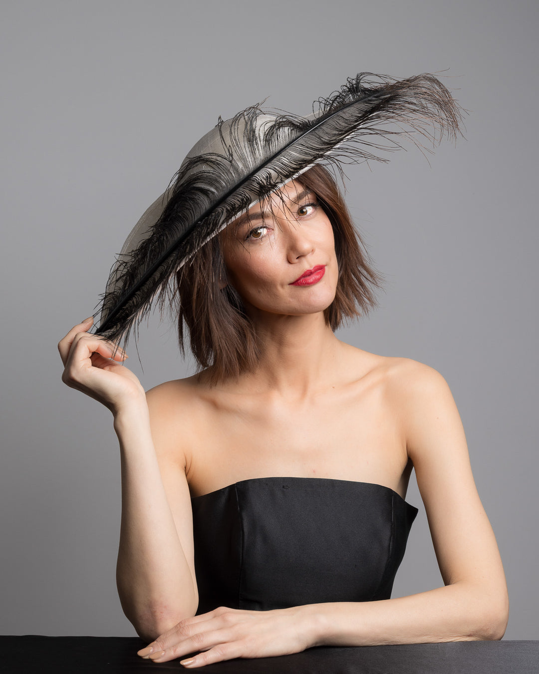 The female model is posing, wearing Kaul, single long, curved feather in shades of black, with fine wispy strands and a visible central quill, placed against a plain white background. Designed and made by Merve Bayindir Millinery
