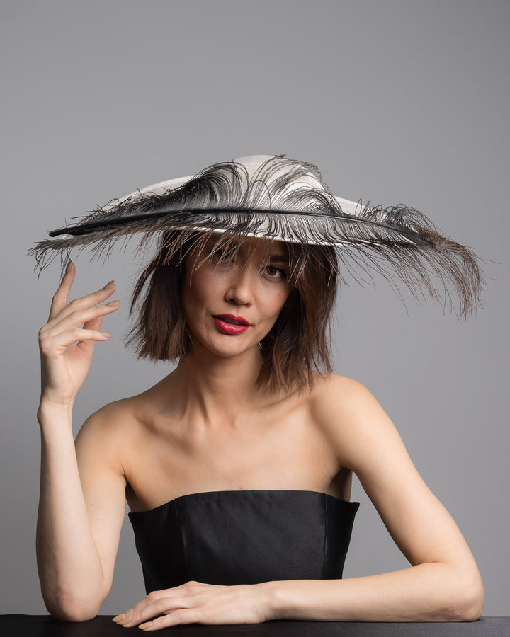 The female model is posing, wearing Kaul, single long, curved feather in shades of black, with fine wispy strands and a visible central quill, placed against a plain white background. Designed and made by Merve Bayindir Millinery