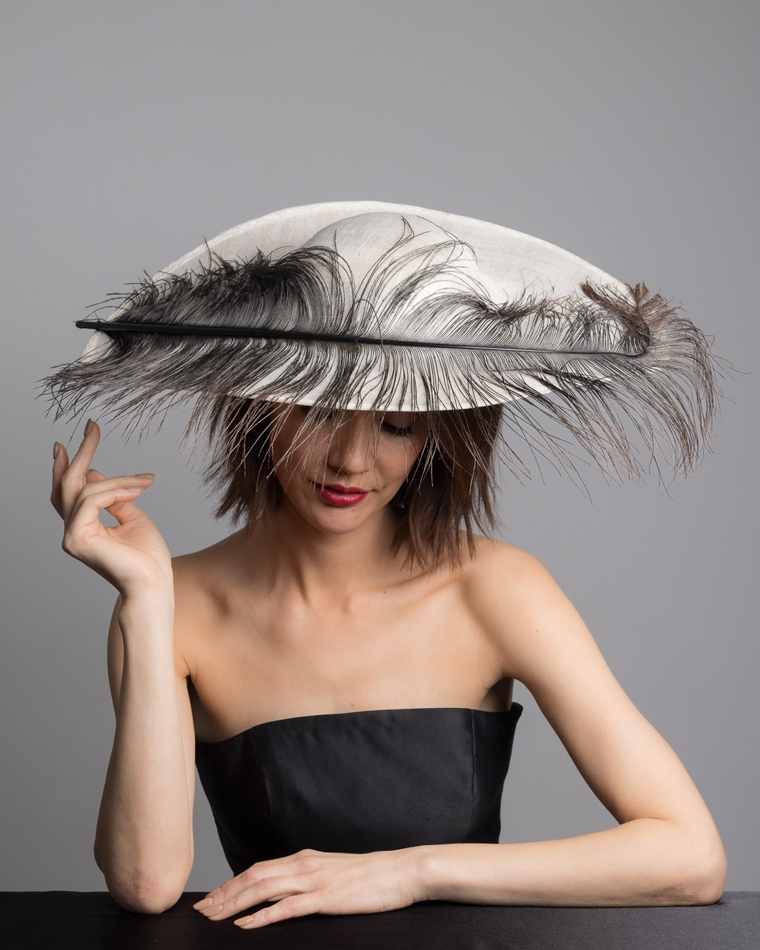 The female model is posing, wearing Kaul, single long, curved feather in shades of black, with fine wispy strands and a visible central quill, placed against a plain white background. Designed and made by Merve Bayindir Millinery