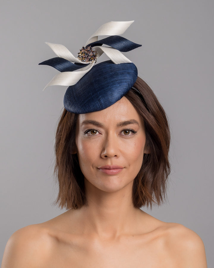 Model is showcasing the front side close up of navy, button shaped fascinator worn to the right side of the head. This chic headpiece is decorated with navy and white buntal swirls joined by multi coloured crystal beads, creating a flower-like silhouette. Designed and made by Merve Bayindir millinery.