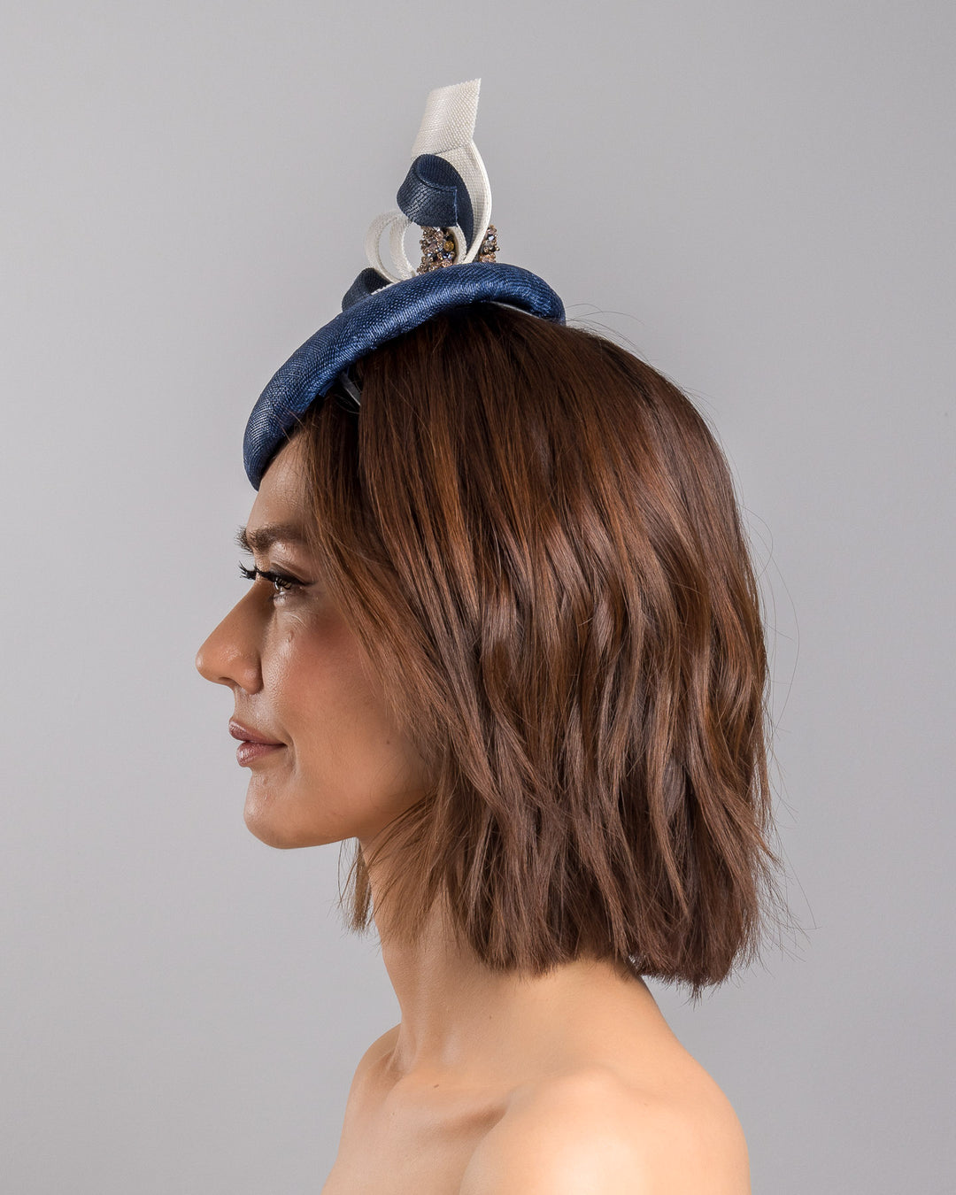 Model is showcasing the left side close up of navy, button shaped fascinator worn to the right side of the head. This chic headpiece is decorated with navy and white buntal swirls joined by multi coloured crystal beads, creating a flower-like silhouette. Designed and made by Merve Bayindir millinery.