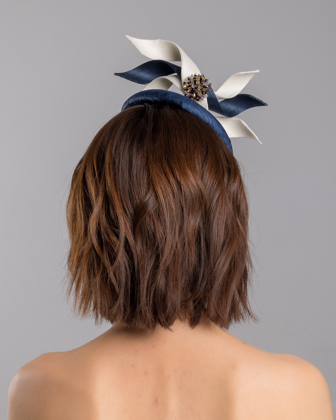 Model is showcasing the back side close up of navy, button shaped fascinator worn to the right side of the head. This chic headpiece is decorated with navy and white buntal swirls joined by multi coloured crystal beads, creating a flower-like silhouette. Designed and made by Merve Bayindir millinery.