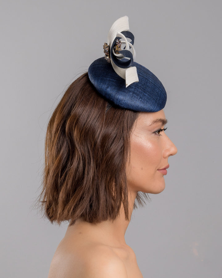 Model is showcasing the right side close up of navy, button shaped fascinator worn to the right side of the head. This chic headpiece is decorated with navy and white buntal swirls joined by multi coloured crystal beads, creating a flower-like silhouette. Designed and made by Merve Bayindir millinery.