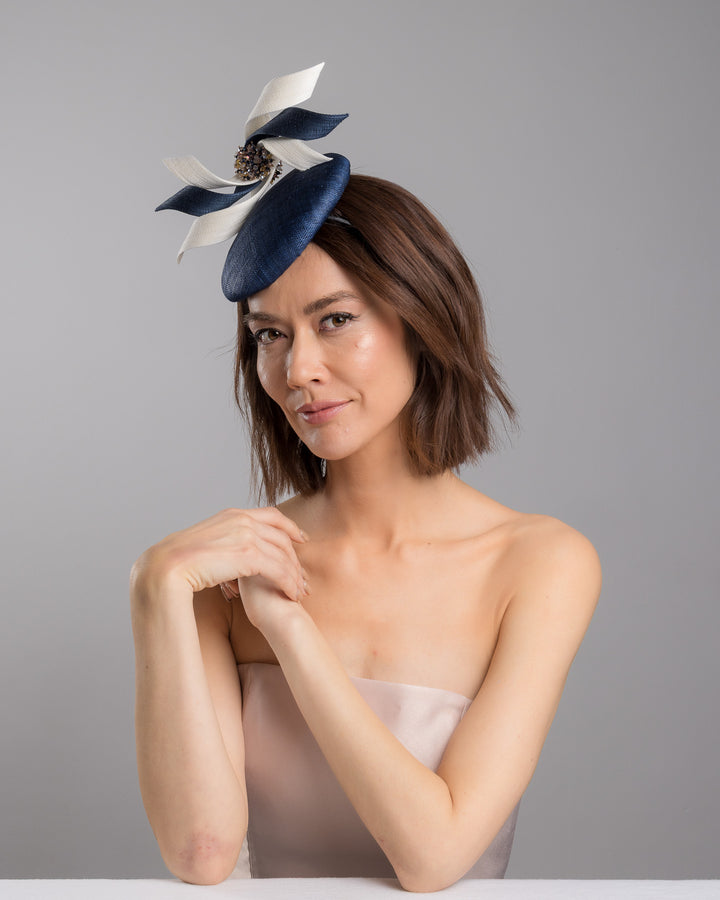 Model is posing wearing Kaden navy button shaped fascinator worn to the right side of the head. This chic headpiece is decorated with navy and white buntal swirls joined by multi coloured crystal beads, creating a flower-like silhouette. Designed and made by Merve Bayindir millinery.
