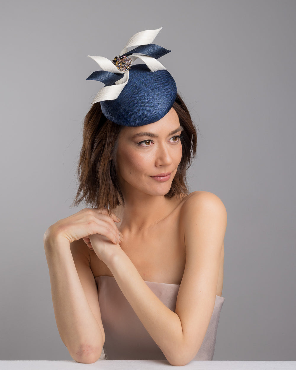 Model is posing wearing Kaden navy button shaped fascinator worn to the right side of the head. This chic headpiece is decorated with navy and white buntal swirls joined by multi coloured crystal beads, creating a flower-like silhouette. Designed and made by Merve Bayindir millinery.