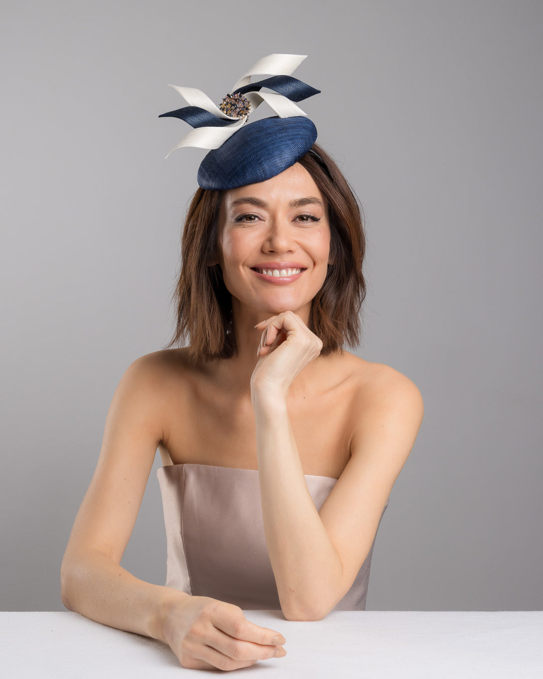 Model is posing wearing Kaden navy button shaped fascinator worn to the right side of the head. This chic headpiece is decorated with navy and white buntal swirls joined by multi coloured crystal beads, creating a flower-like silhouette. Designed and made by Merve Bayindir millinery.