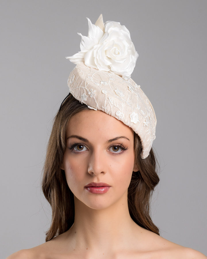 The model is showcasing the front side close up of the light pink, percher style sinamay hat that curves on the left side of the head, covered with flower detailed white lace. This pink fascinator features a large, white silk satin rose that enhances the visual impact. Designed and made by Merve Bayindir Millinery.