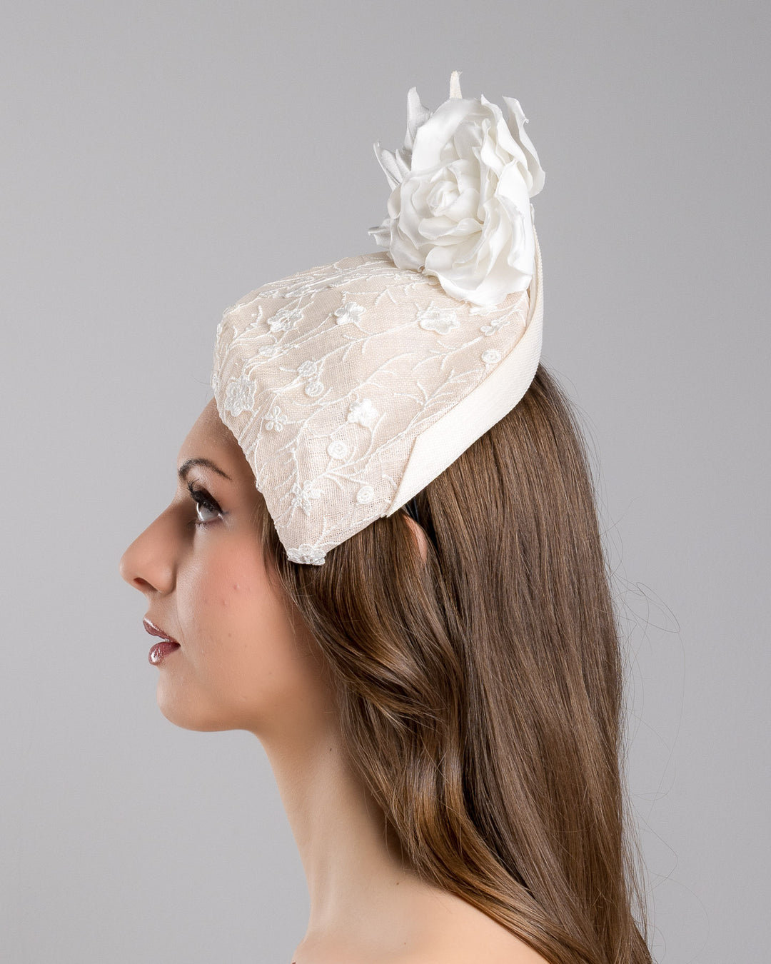 The model is showcasing the left side close up of the light pink, percher style sinamay hat that curves on the left side of the head, covered with flower detailed white lace. This pink fascinator features a large, white silk satin rose that enhances the visual impact. Designed and made by Merve Bayindir Millinery.
