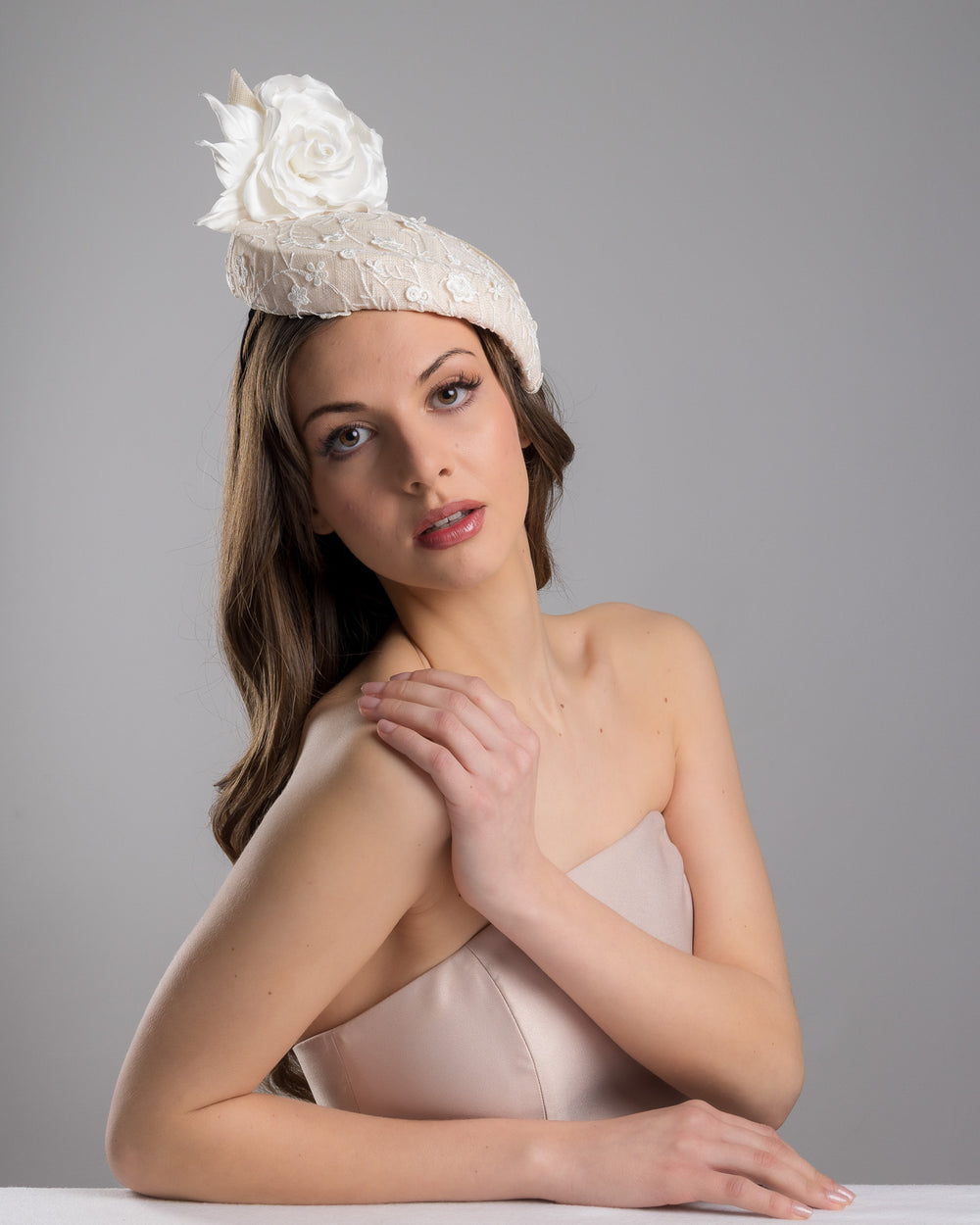 The female model is posing, wearing Kerry,the light pink, percher style sinamay hat that curves on the left side of the head, covered with flower detailed white lace. This pink fascinator features a large, white silk satin rose that enhances the visual impact. Designed and made by Merve Bayindir Millinery.