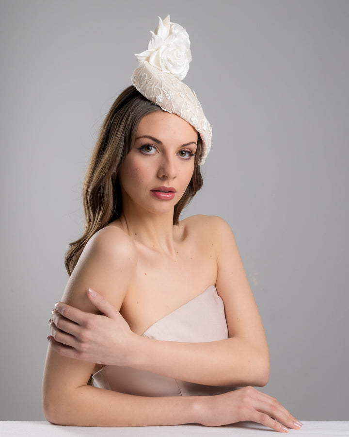 The female model is posing, wearing Kerry,the light pink, percher style sinamay hat that curves on the left side of the head, covered with flower detailed white lace. This pink fascinator features a large, white silk satin rose that enhances the visual impact. Designed and made by Merve Bayindir Millinery.