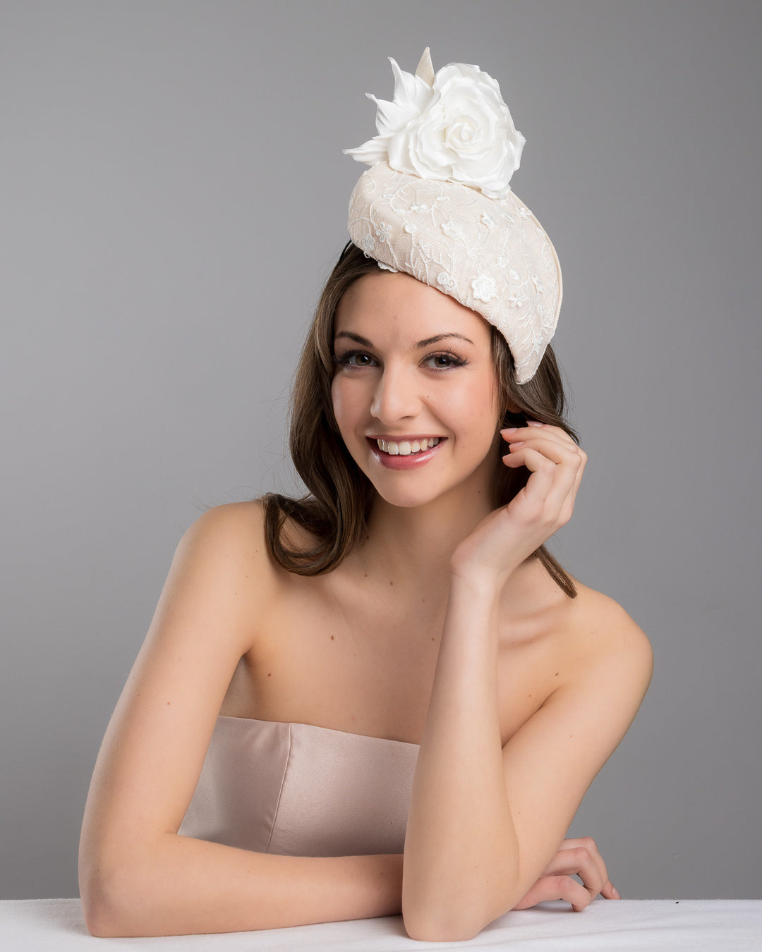 The female model is posing, wearing Kerry,the light pink, percher style sinamay hat that curves on the left side of the head, covered with flower detailed white lace. This pink fascinator features a large, white silk satin rose that enhances the visual impact. Designed and made by Merve Bayindir Millinery.