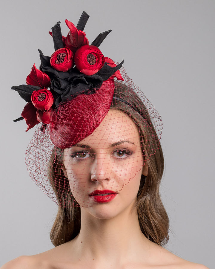 Model is showcasing the front side close up of red button shaped fascinator worn to the centre to the right side of the head. This fascinator hat is decorated with a bouquet of black crepe roses, black shaded red peonies and black buntal swirls, and, rounded with a delicate red veil that covers the face. Designed and made by Merve Bayindir millinery.