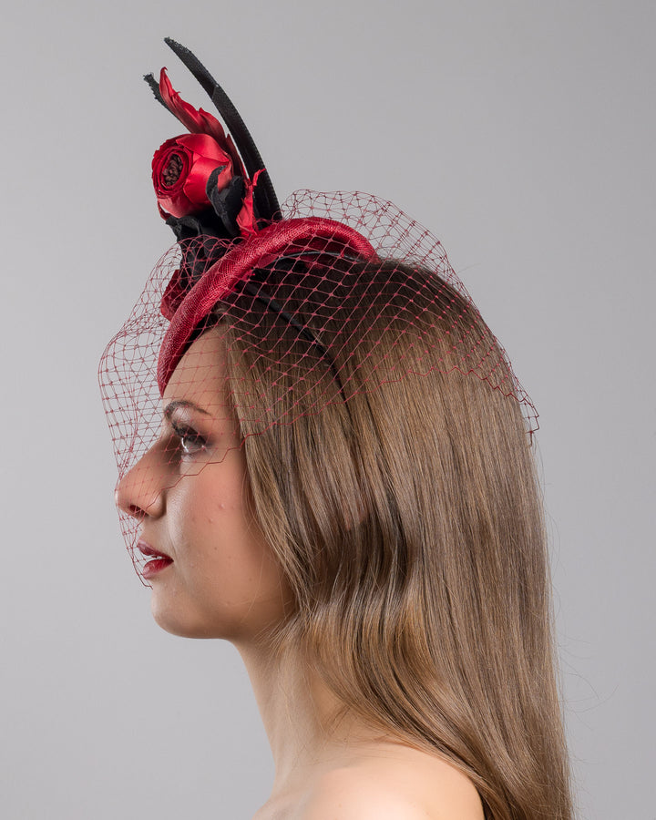 Model is showcasing the left side close up of red button shaped fascinator worn to the centre to the right side of the head. This fascinator hat is decorated with a bouquet of black crepe roses, black shaded red peonies and black buntal swirls, and, rounded with a delicate red veil that covers the face. Designed and made by Merve Bayindir millinery.