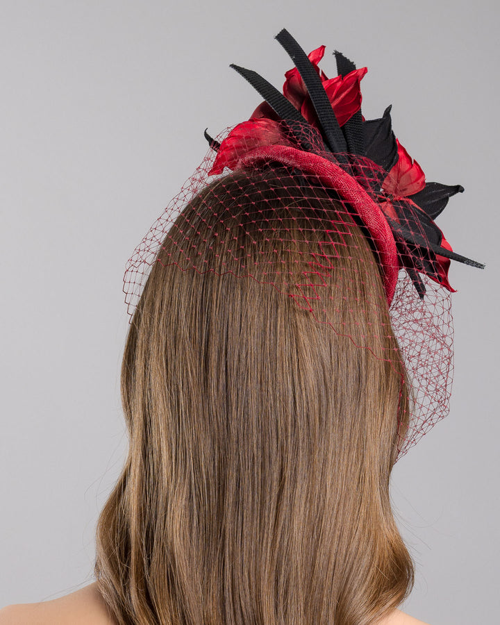 Model is showcasing the back side close up of red button shaped fascinator worn to the centre to the right side of the head. This fascinator hat is decorated with a bouquet of black crepe roses, black shaded red peonies and black buntal swirls, and, rounded with a delicate red veil that covers the face. Designed and made by Merve Bayindir millinery.