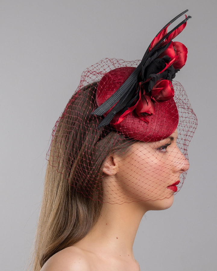 Model is showcasing the right side close up of red button shaped fascinator worn to the centre to the right side of the head. This fascinator hat is decorated with a bouquet of black crepe roses, black shaded red peonies and black buntal swirls, and, rounded with a delicate red veil that covers the face. Designed and made by Merve Bayindir millinery.