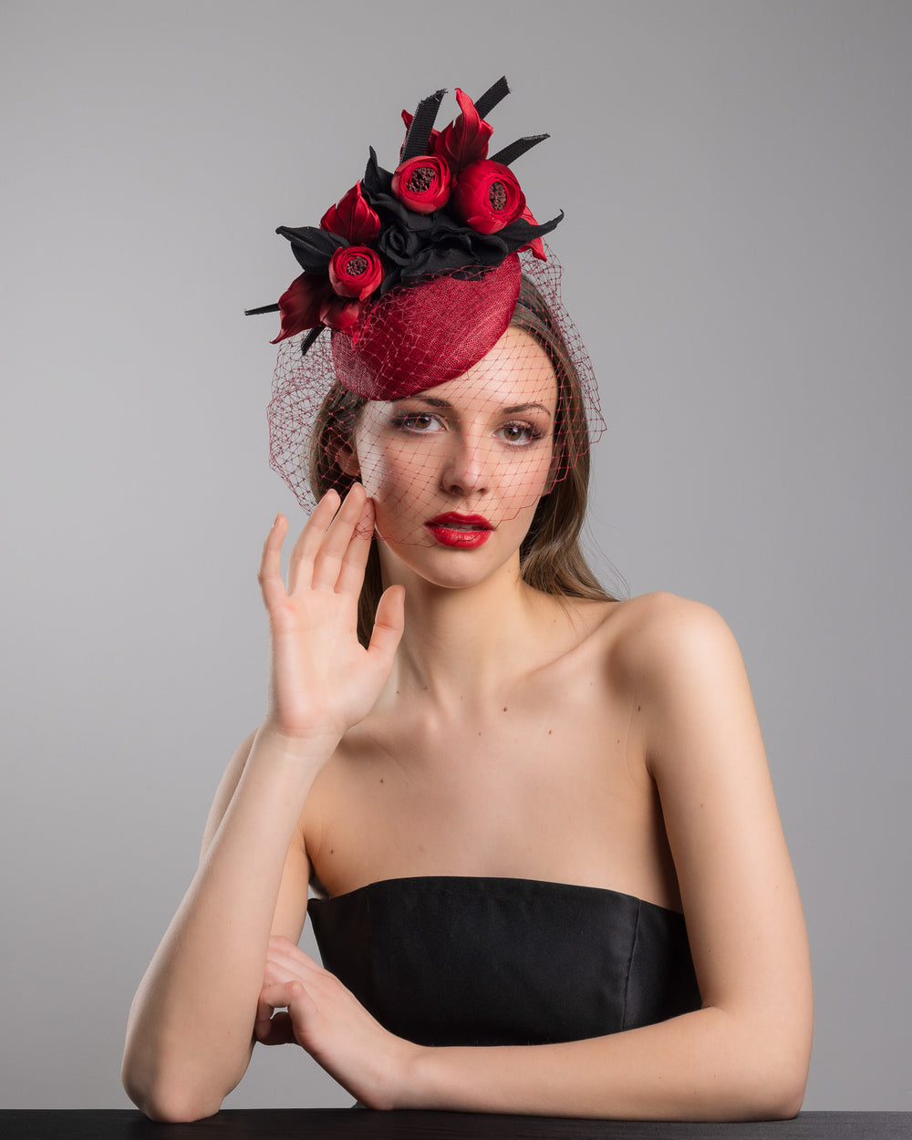 Female model is posing wearing Kingsley the red button shaped fascinator worn to the centre to the right side of the head. This fascinator hat is decorated with a bouquet of black crepe roses, black shaded red peonies and black buntal swirls, and, rounded with a delicate red veil that covers the face. Designed and made by Merve Bayindir millinery.