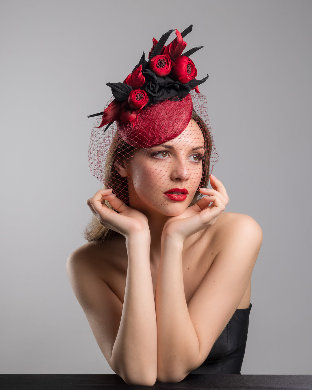 Female model is posing wearing Kingsley the red button shaped fascinator worn to the centre to the right side of the head. This fascinator hat is decorated with a bouquet of black crepe roses, black shaded red peonies and black buntal swirls, and, rounded with a delicate red veil that covers the face. Designed and made by Merve Bayindir millinery.