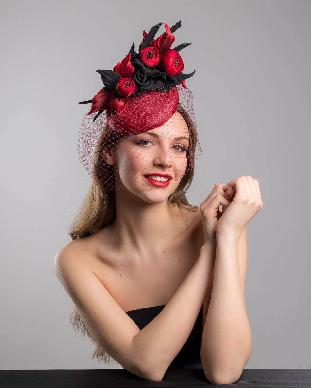 Female model is posing wearing Kingsley the red button shaped fascinator worn to the centre to the right side of the head. This fascinator hat is decorated with a bouquet of black crepe roses, black shaded red peonies and black buntal swirls, and, rounded with a delicate red veil that covers the face. Designed and made by Merve Bayindir millinery.