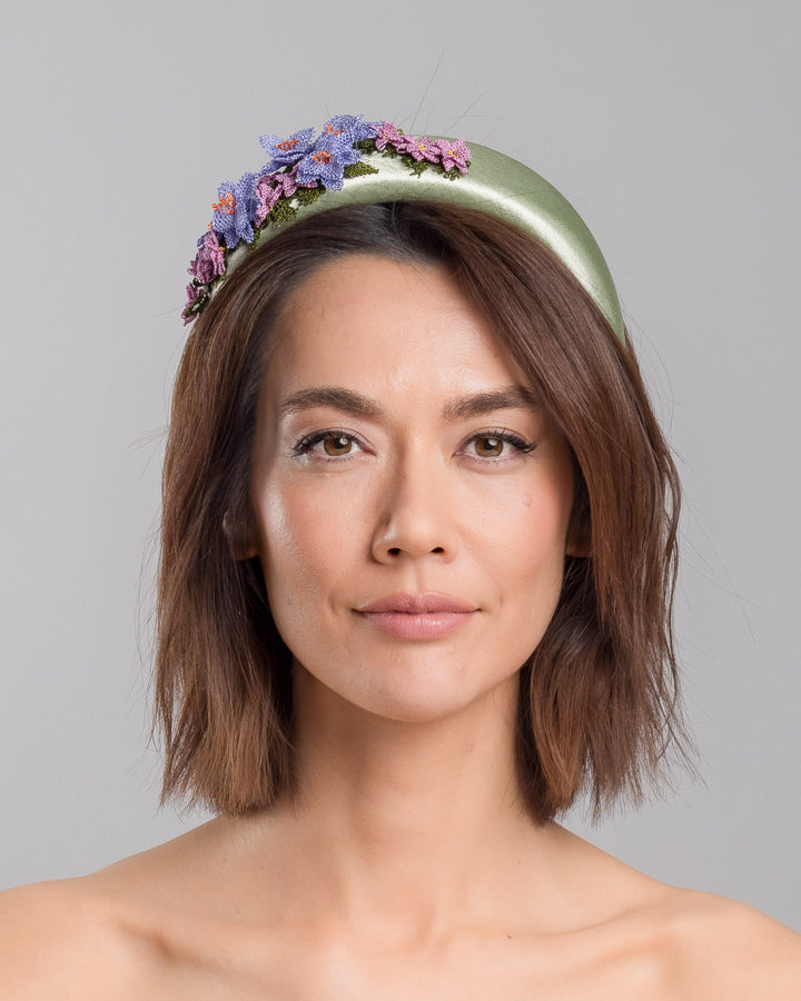 The model is showcasing the front side close up of the sage green satin padded headband embellished with handcrafted Turkish needle embroidery flowers in lilac and pink tones, featuring detailed petals and green stitched leaves clustered along the top of the band.  Designed and made by Merve Bayindir Millinery.
