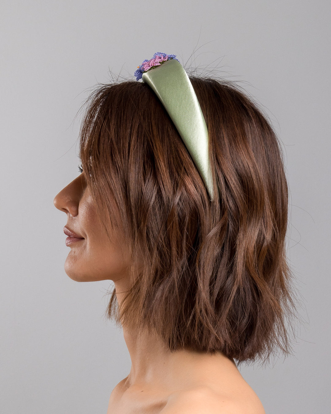 The model is showcasing the left side close up of the sage green satin padded headband embellished with handcrafted Turkish needle embroidery flowers in lilac and pink tones, featuring detailed petals and green stitched leaves clustered along the top of the band.  Designed and made by Merve Bayindir Millinery.
